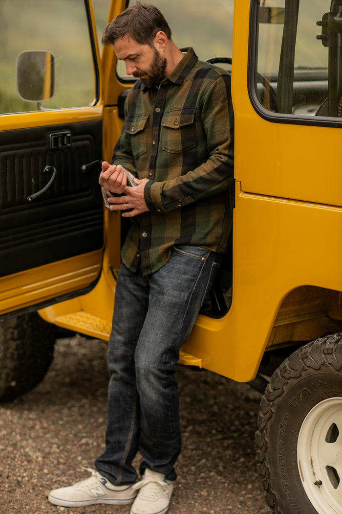 A man in a Pladra Men's Fireside Flannel - Woodland Green and jeans stands by an open yellow vehicle door, looking down.