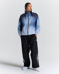 A man modeling a light blue and navy gradient zip-up jacket with black track pants featuring light blue star details, set against a plain light gray studio background.