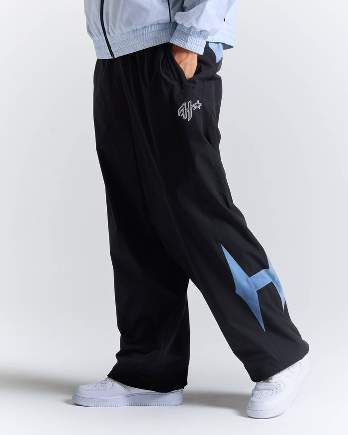 A person wearing black wide-leg sweatpants with a light blue graphic on the lower leg, paired with a light blue zip jacket and white sneakers, shown from the waist down against a light gray studio background.
