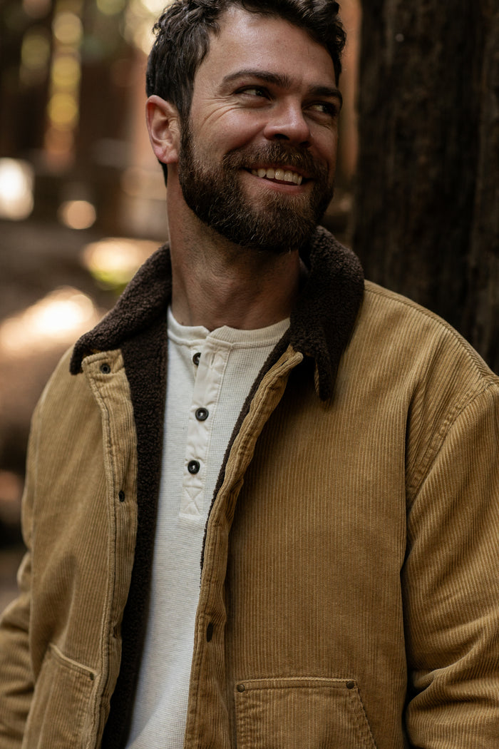 A bearded man in a Pladra Men's Ironwood Sherpa Jacket - Acorn Tan smiles outdoors in a forest.
