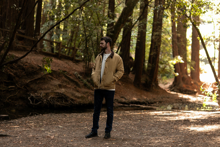 A man wears the Pladra Men's Ironwood Sherpa Jacket in Acorn Tan, standing in a sunlit forest with hands in his pockets.