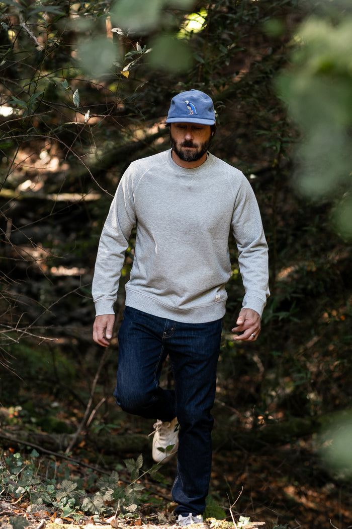 A man in a blue cap, jeans, and the Men's Klamath Crew - Fog Grey walks through a sunlit wooded area.