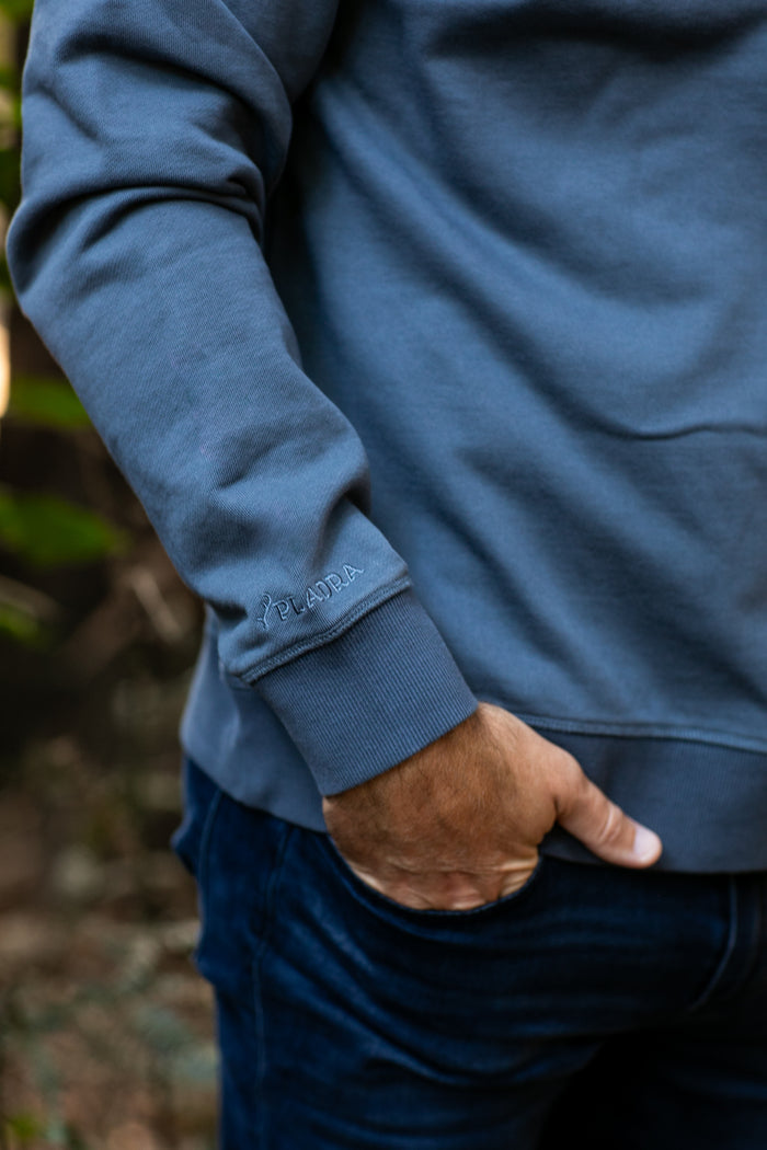 Close-up of a man's arm in Pladra Men's Klamath Crew - Stone Blue, logo on sleeve, hand in pocket.
