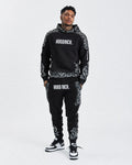 A man modeling a black and white patterned hoodie and matching sweatpants with 'HOODRICH.' printed in white on the chest and thighs, paired with black and white sneakers, against a light gray background.