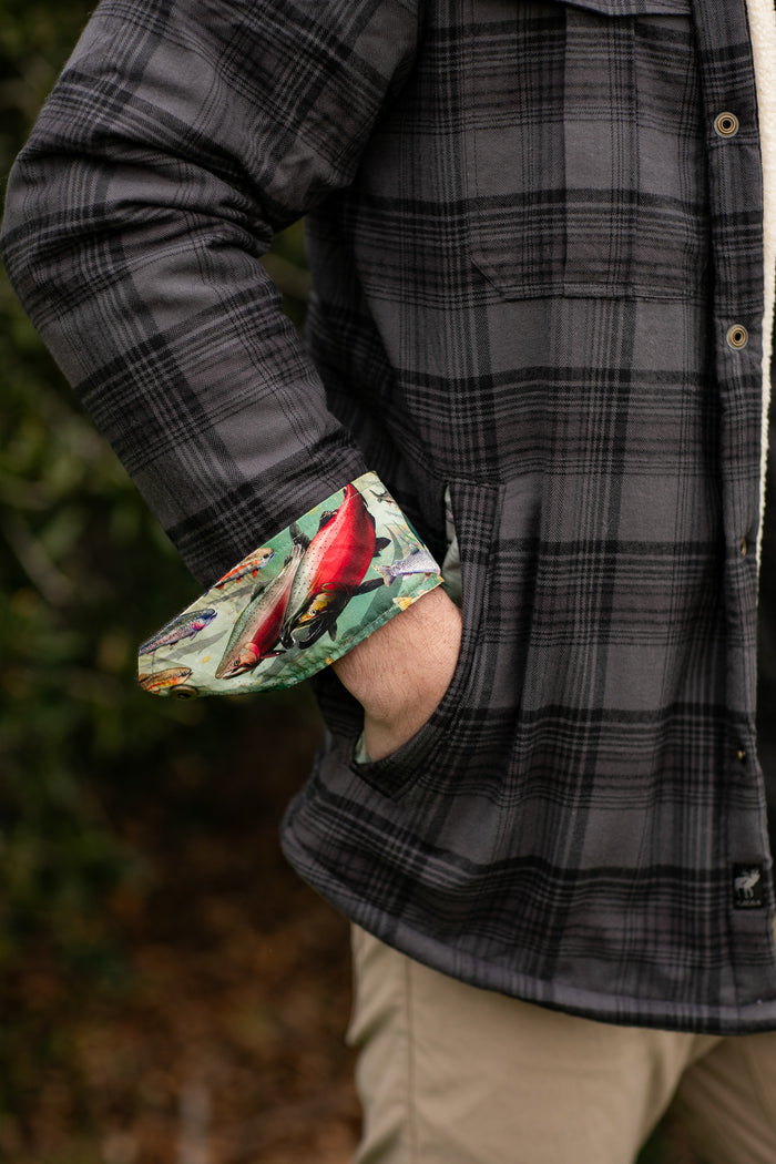 Person wearing a Pladra Men's Northwood Sherpa Insulated Flannel Jacket in Deadwood Black, hand in pocket, outdoors.