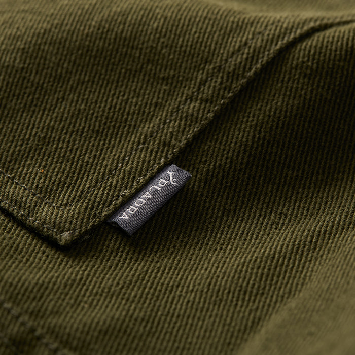 Close-up of hunter green cotton fabric with a visible seam and a small black Pladra tag on the Men's Sequoia Chore Coat.
