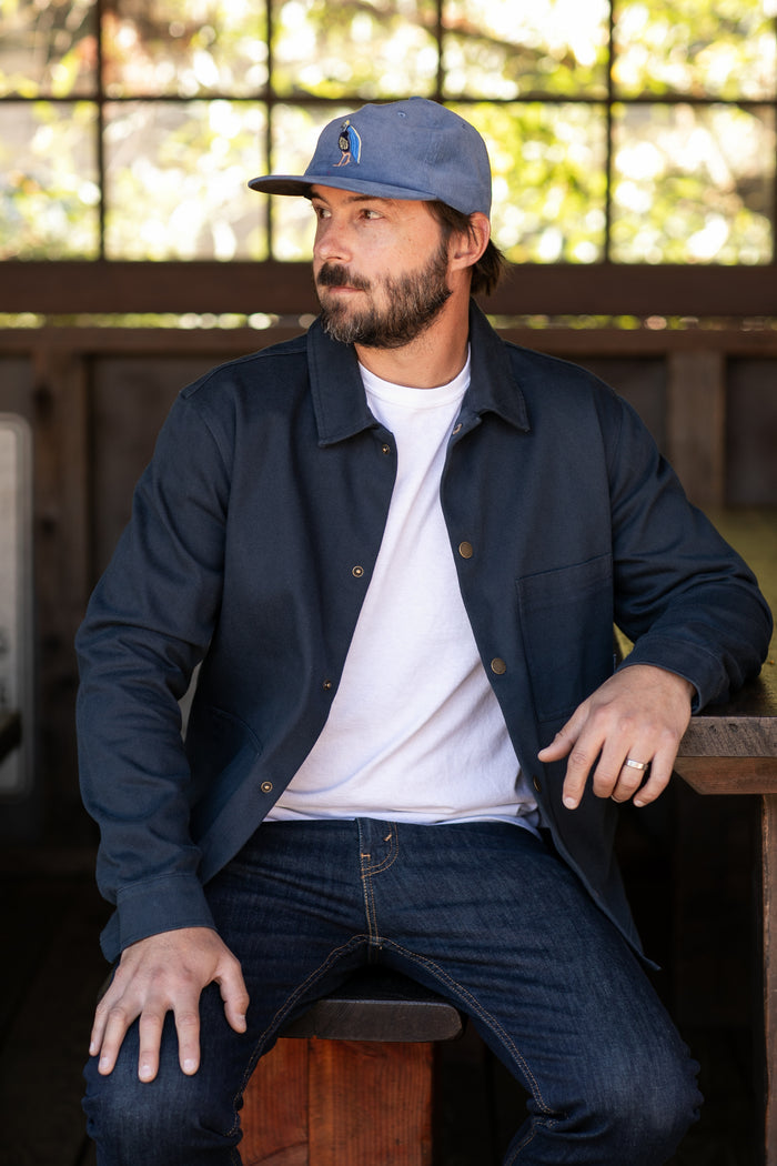 A bearded man in a blue cap, Men's Sequoia Chore Coat - Midnight Blue, white t-shirt, and jeans sits on a stool indoors by a wooden table, looking left as sunlight streams through a large window behind him.