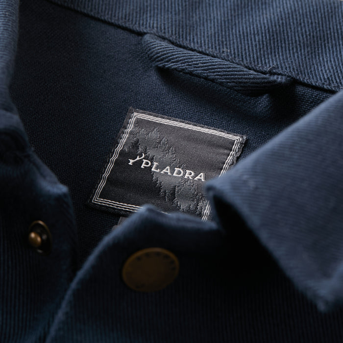 Close-up of the Men's Sequoia Chore Coat in Midnight Blue, styled like a cotton twill jacket with a Pladra label featuring a tree design inside. Brass buttons near the collar add a classic workwear touch.