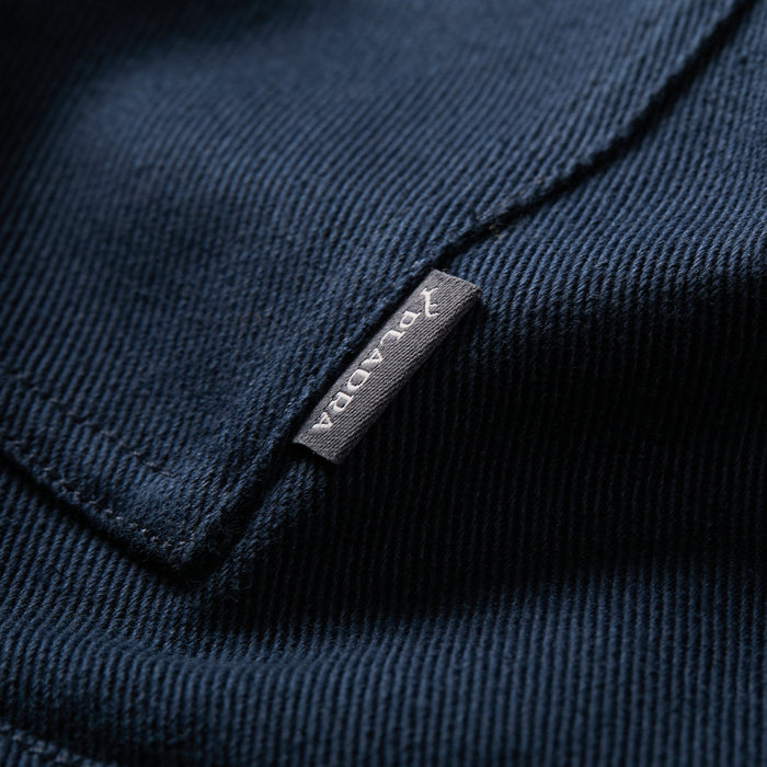 Close-up of the Men's Sequoia Chore Coat in Midnight Blue, a ribbed cotton twill jacket with a small gray tag reading JAVADAR in white, blending classic workwear style and modern details.