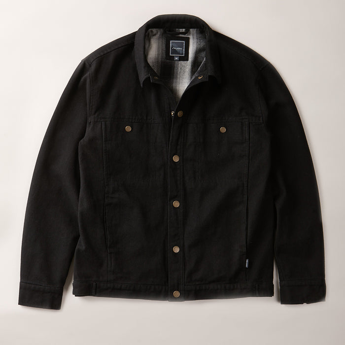 The Men's Sequoia Jacket- Shadow Black, made from black cotton twill with metal buttons, chest flap pockets, and side pockets, is laid flat on a light-colored surface.