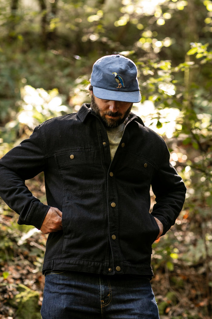 A man in jeans and a blue baseball cap stands outdoors in a sunlit forest, looking down with his hands in the pockets of his Men's Sequoia Jacket - Shadow Black.