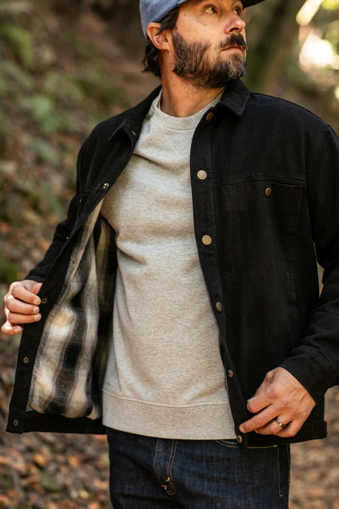 A bearded man in a gray cap and sweatshirt stands outdoors, showing the plaid flannel lining inside his open Men's Sequoia Jacket- Shadow Black as he looks up and adjusts the cotton twill jacket with one hand.
