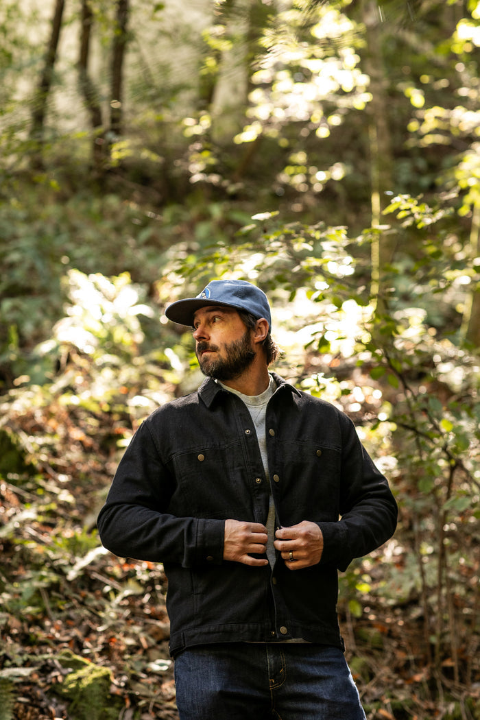 A man in jeans and a blue cap stands in a sunlit forest, buttoning his Men's Sequoia Jacket - Shadow Black. Sunlight filters through the trees, highlighting the flannel-lined cotton twill jacket and the lush greenery around him.