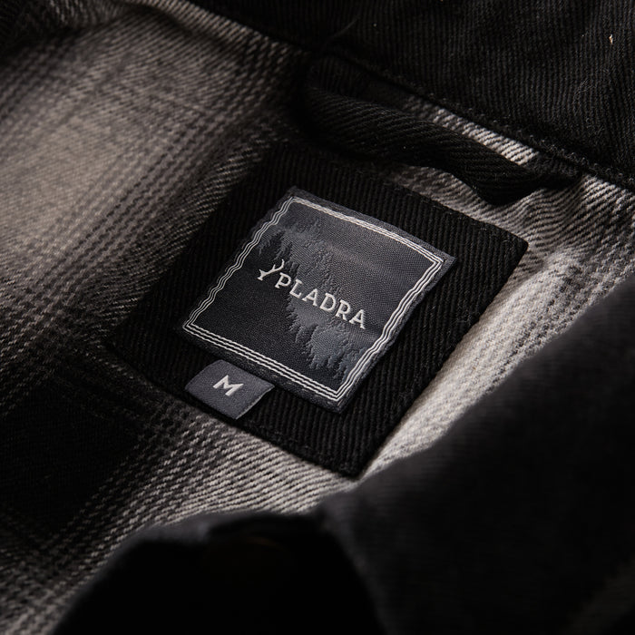 A close-up of the Men's Sequoia Jacket- Shadow Black, a black and gray plaid cotton twill jacket featuring a Pledra label, size M tag, and classic flannel-lined details.