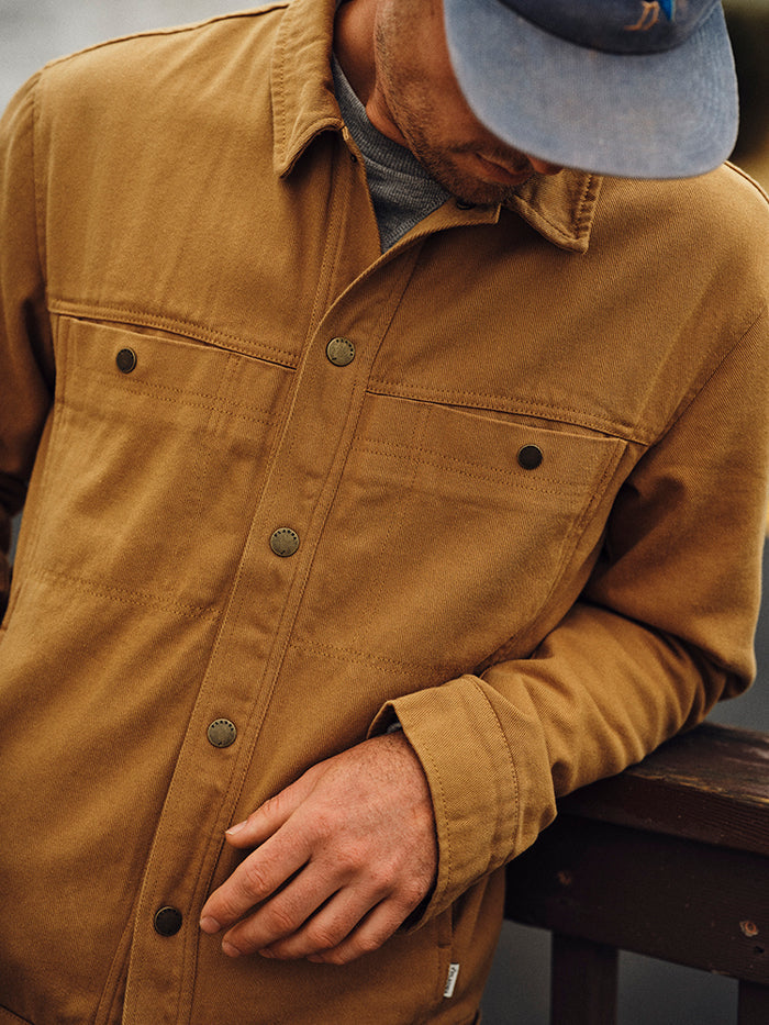 Man in a blue cap looks down, resting his arm on a railing, wearing a Pladra Men's Sequoia Jacket in Tobacco Brown.