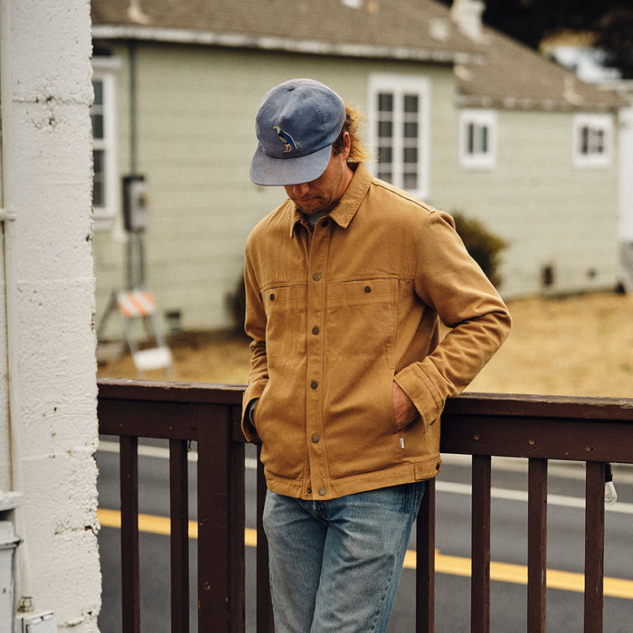 A man in a Pladra Men's Sequoia Jacket - Tobacco Brown and blue cap stands on a wooden porch, hands in pockets.