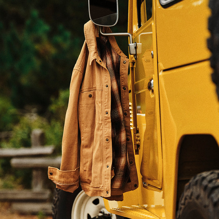 A Pladra Men's Sequoia Jacket in Tobacco Brown hangs on a yellow vehicle’s mirror, trees and fence behind.