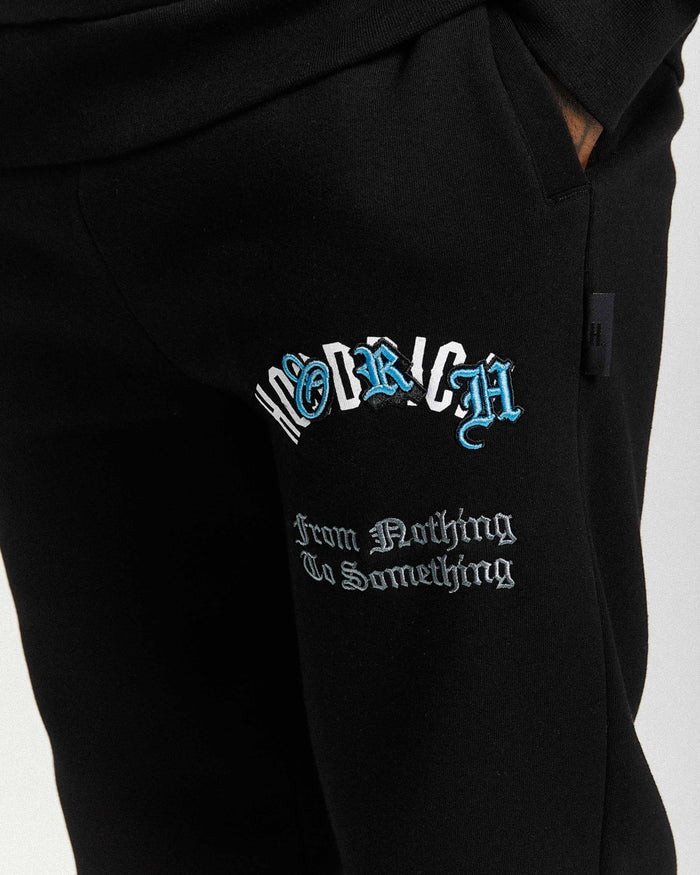 Close-up of black sweatpants with a drawstring waist, featuring a stylized 'Hoodrich' logo in white and blue gothic font and the phrase 'From Nothing To Something' in white gothic text on the left thigh.