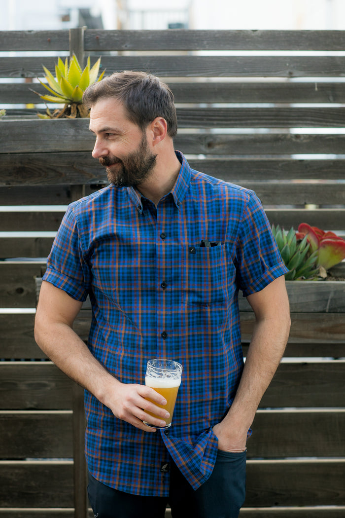 A man wears the Pladra Men's Short Sleeve Tailwinds Shirt in Lagoon Blue, holding a beer by a wooden fence with plants.