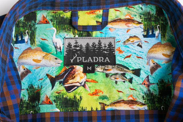 A fabric tag on a Pladra Men's Short Sleeve Tailwinds Shirt - Lagoon Blue with fish, birds, and trees around the label.