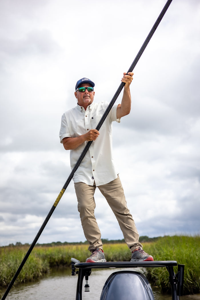 Wearing the Men's Short Sleeve Tailwinds Shirt in Sand White Chambray and khaki pants, a man in sunglasses stands at the front of a boat, guiding it through a grassy marsh with a long pole beneath a cloudy sky.