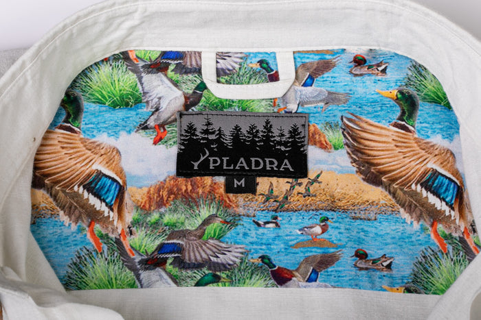 The Men's Short Sleeve Tailwinds Shirt - Sand White Chambray features a soft cotton chambray build and an inside collar with a colorful duck pattern near grass and water, plus a centered black PLADRA M label. Lightweight with an active fit.