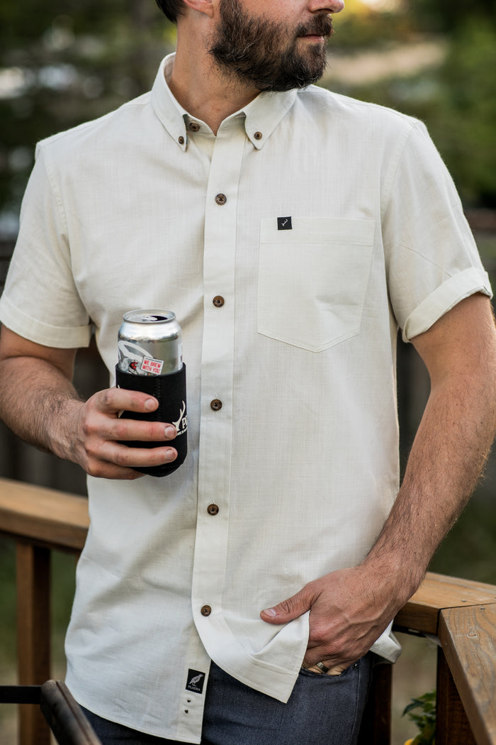 A man stands outdoors by a wooden railing, holding a canned drink with a black koozie and wearing the Men's Short Sleeve Tailwinds Shirt in Sand White Chambray, with his other hand resting in his pocket.