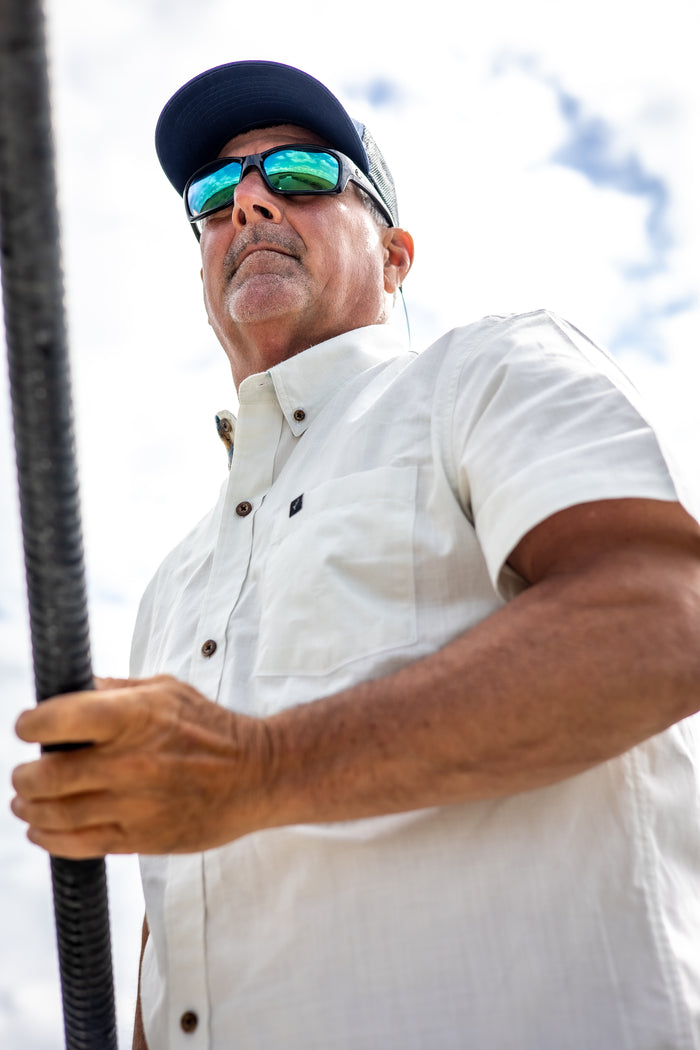 A man in sunglasses, a cap, and the Men's Short Sleeve Tailwinds Shirt - Sand White Chambray stands outdoors with a rod against a cloudy sky, looking ahead seriously.