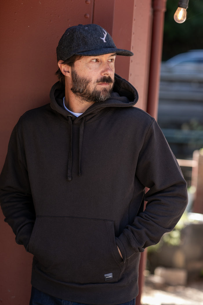 A man wears the Pladra Men's Sierra Hoodie in Charcoal Black and a cap, standing outdoors with hands in his pockets.
