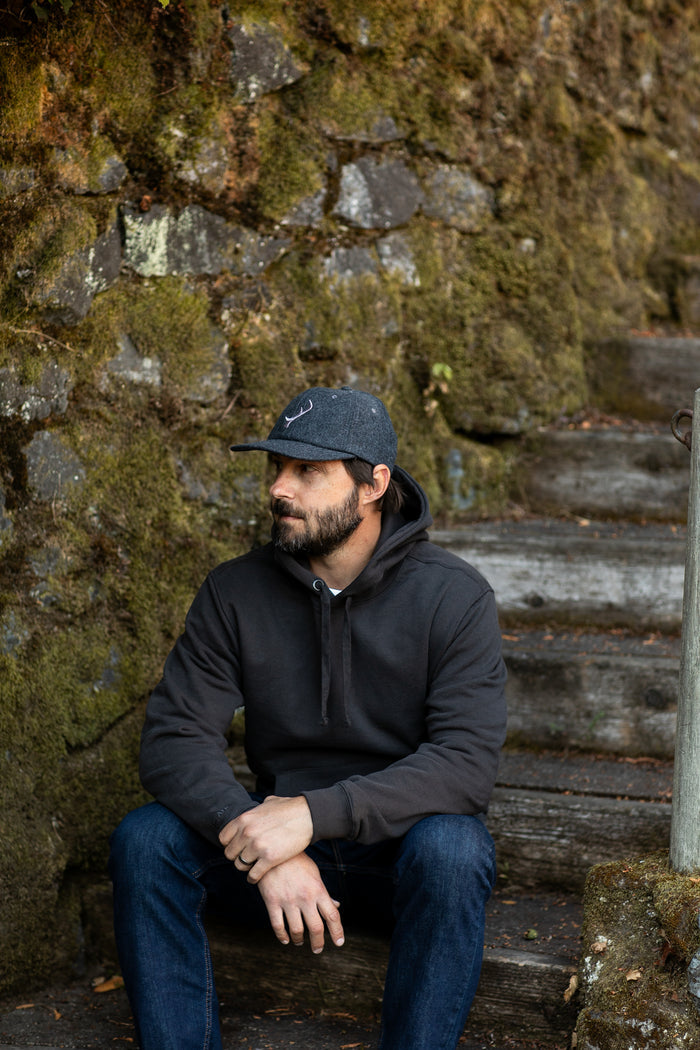 Man wears Pladra Men's Sierra Hoodie—Charcoal Black, sitting relaxed on stone steps by a mossy wall, looking left.
