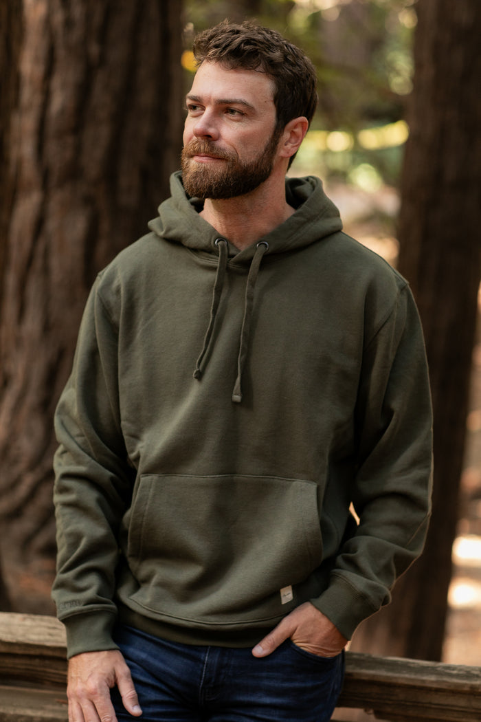 A bearded man wearing the Men's Sierra Hoodie in Pine Green and blue jeans stands outdoors by a wooden railing, trees and soft sunlight in the background.