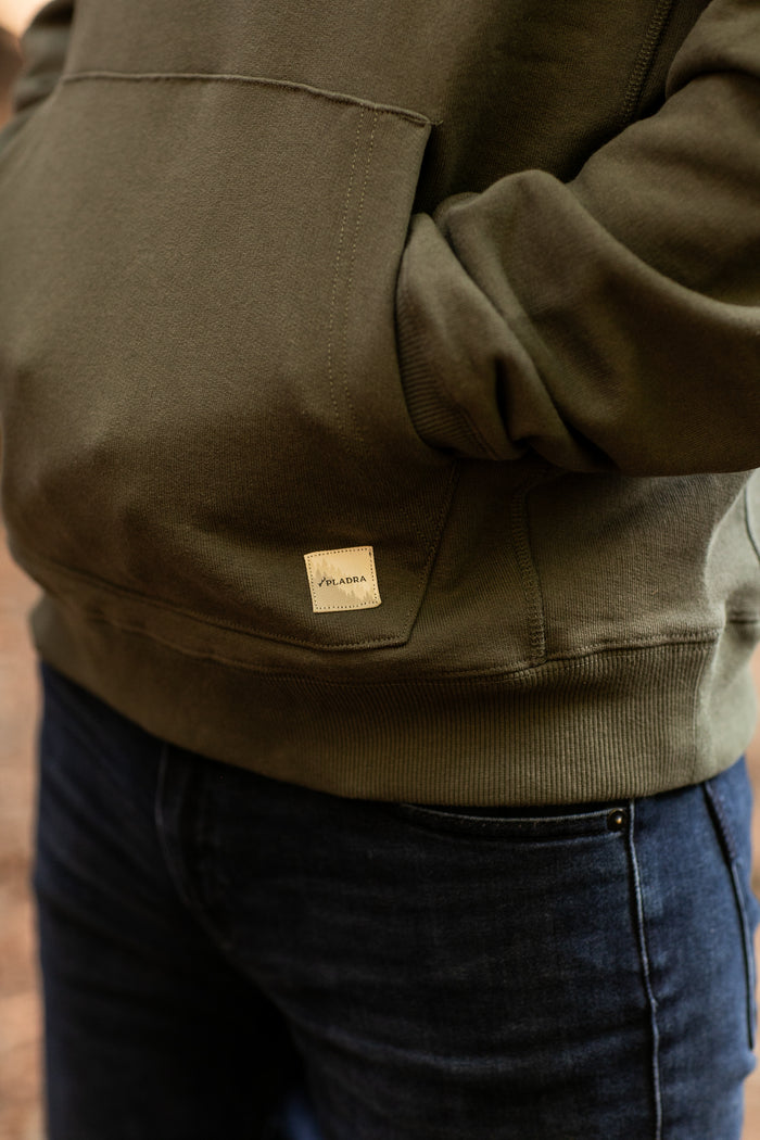 A person wearing the Men's Sierra Hoodie-Pine Green with hands in the front pocket and dark blue jeans. A small beige tag with text is visible on this premium cotton hoodie near the pocket.