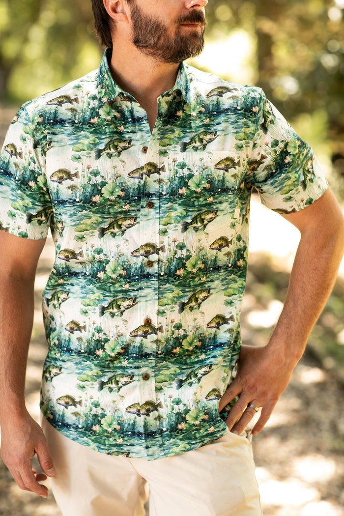 Man wearing a Pladra Men's S/S Printed Outdoor Aloha Shirt - Bass and beige pants, standing outdoors with trees.