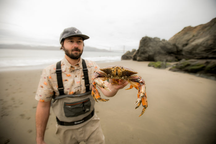 A man in a Pladra Men's S/S Printed Outdoor Aloha Shirt holds a large crab on the beach with rocks and ocean behind.