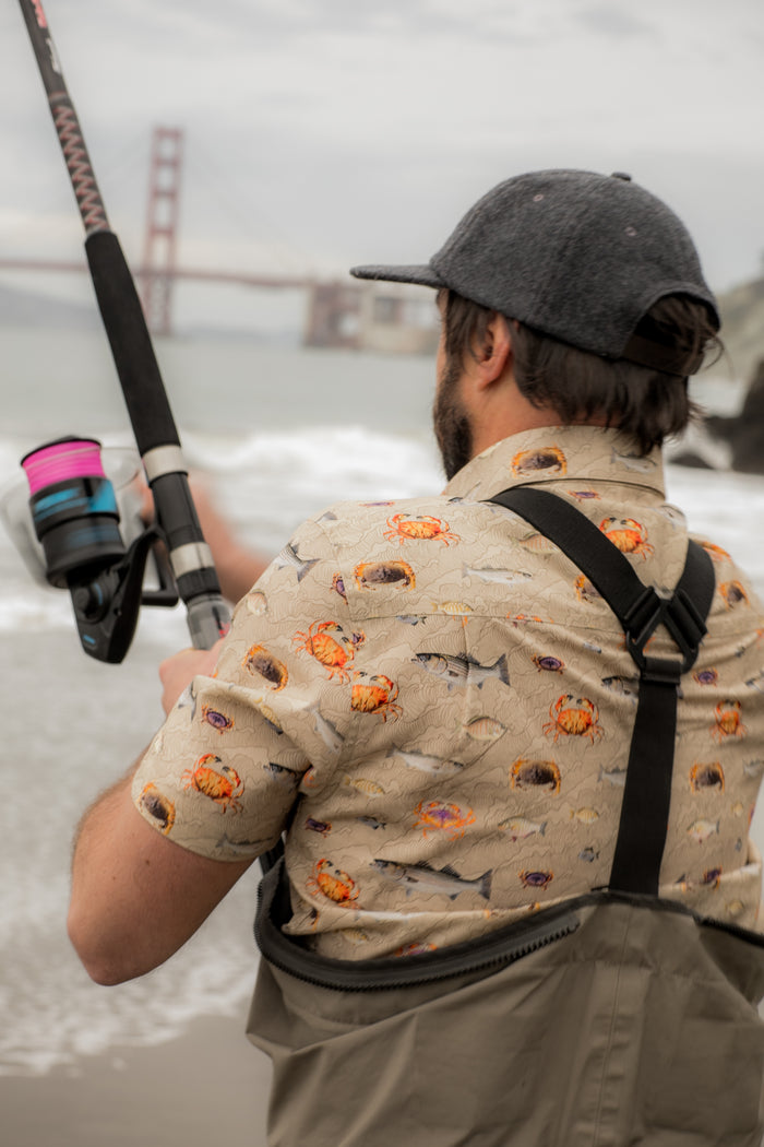 Wearing Pladra's Men's S/S Printed Outdoor Aloha Shirt - Bay Surf, a man fishes by the ocean near the Golden Gate Bridge.
