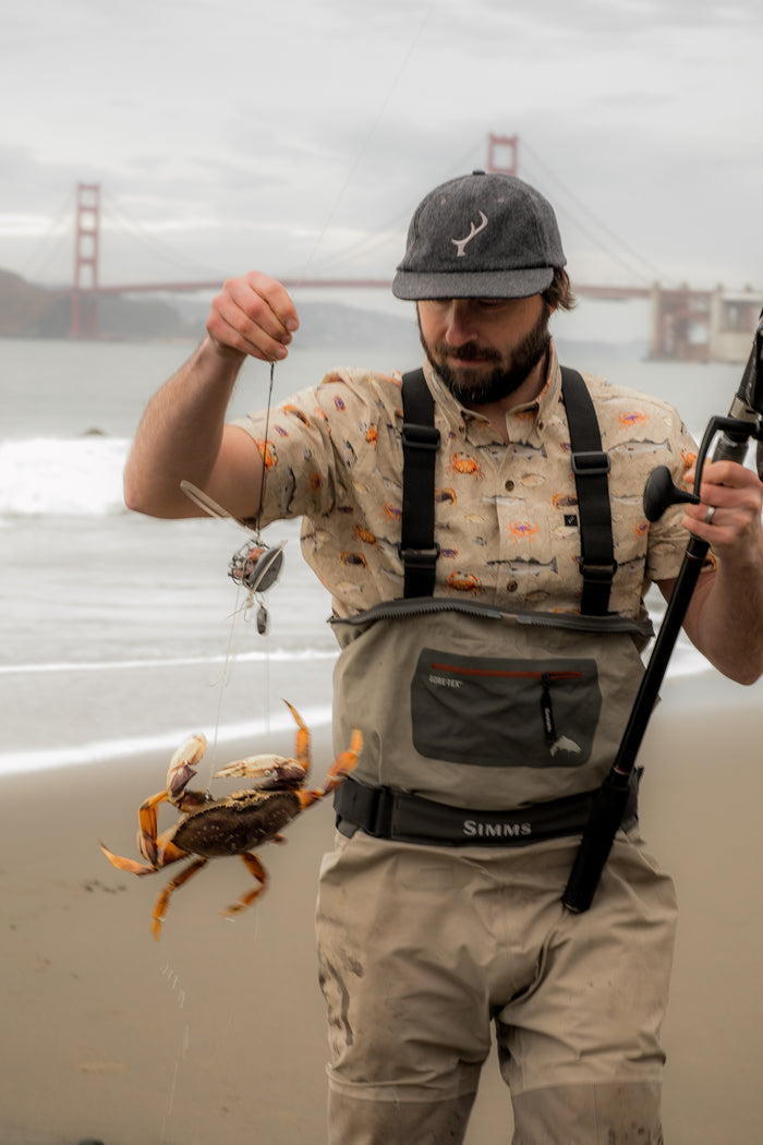 A man in a Pladra Men's S/S Printed Outdoor Aloha Shirt - Bay Surf holds a fishing rod and crab on the beach.