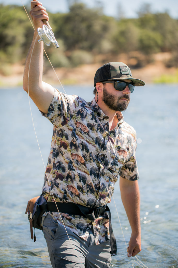 Man in sunglasses wears Pladra Men's S/S Printed Outdoor Aloha Shirt - Bears, fly fishing with a rod in a shimmering river.