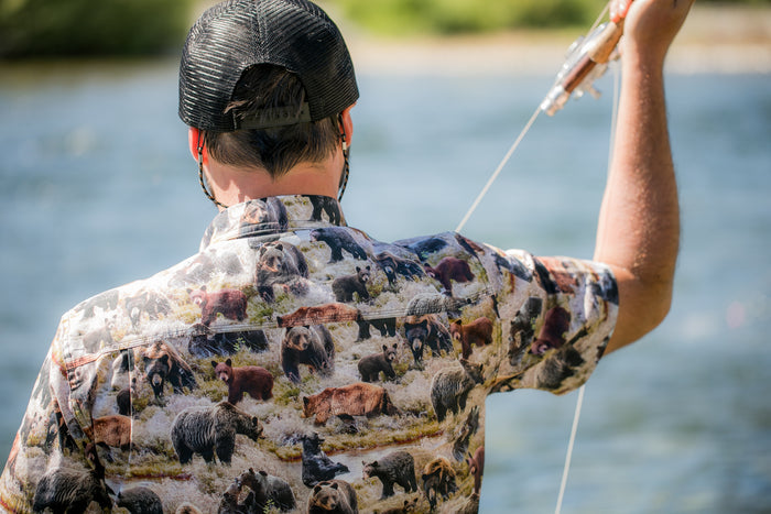 Man fly-fishing in a Pladra Men's S/S Printed Outdoor Aloha Shirt - Bears, by a sunny riverbank, seen from behind.