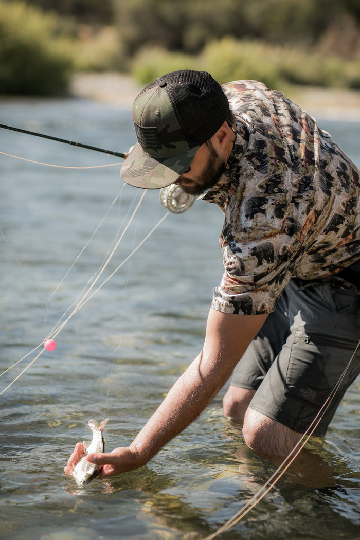 A man in a Pladra Men's S/S Printed Outdoor Aloha Shirt - Bears catches a fish fly fishing in a clear, shallow river.