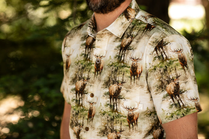 A person wears the Men's S/S Printed Outdoor Aloha Shirt - Elk, featuring a short-sleeved cotton design with illustrated elk in a forest. The background displays blurred green foliage, and the person’s face is partly out of frame.
