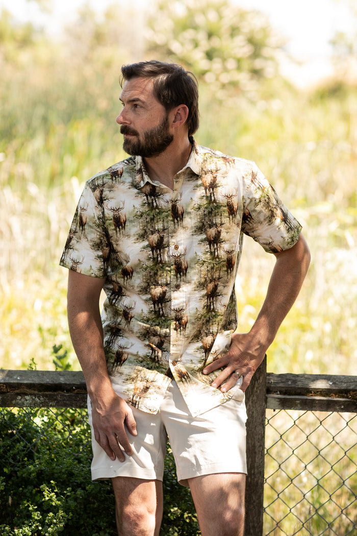 A man with short dark hair and a beard stands by a wooden fence outdoors, wearing the Men's S/S Printed Outdoor Aloha Shirt - Elk and light shorts, with lush greenery in the sunlit, blurred background.
