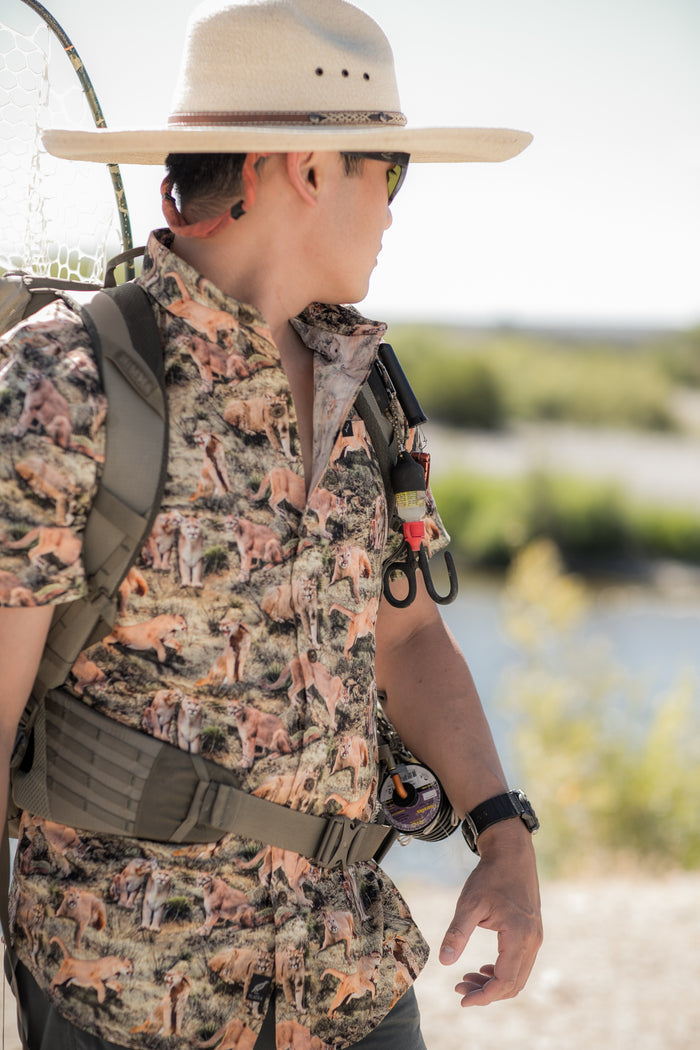 A person in a wide-brimmed hat, sunglasses, and backpack stands outdoors wearing the Men's S/S Printed Outdoor Aloha Shirt - Mountain Lions, featuring vibrant prints. They look to the side with blurred greenery and water behind them.