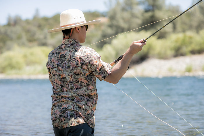 Wearing a wide-brimmed hat, sunglasses, and the Men's S/S Printed Outdoor Aloha Shirt - Mountain Lions, a person fly fishes in a sunny river surrounded by lush trees and greenery.
