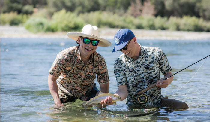 Two men kneel in shallow water fishing; one with a rod, the other holding a fish. Both wear Men's S/S Printed Outdoor Aloha Shirt - Mountain Lions and hats, smiling with trees and a riverbank in the background.