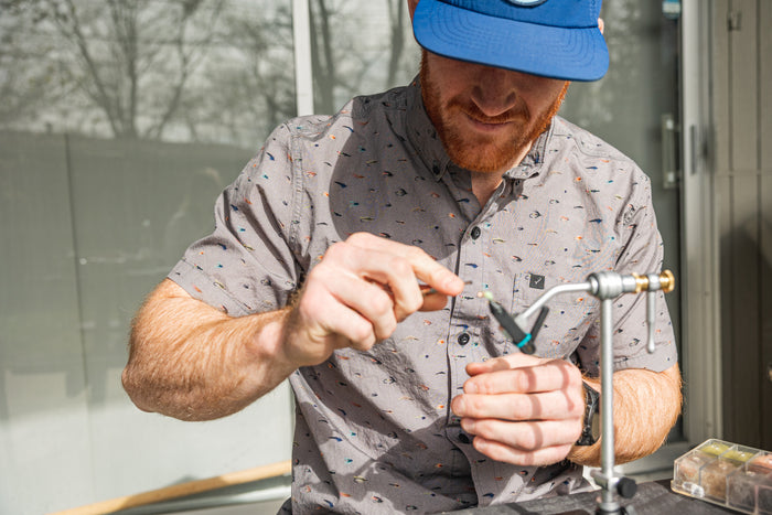 A man in a Pladra Men's S/S Printed Outdoor Aloha Shirt ties a fishing fly on a sunny patio.