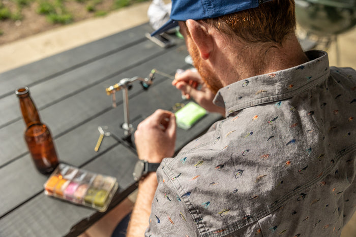 Wearing the Pladra Men's S/S Printed Outdoor Aloha Shirt - Steelhead Flies, he ties fishing flies outdoors with a beer.