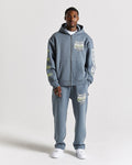 A man modeling a matching slate blue zip-up hoodie and sweatpants set with white sneakers, photographed against a neutral light gray studio background.
