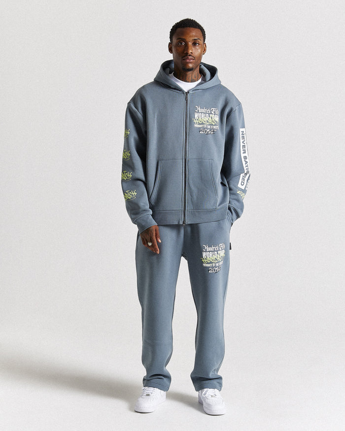 A man modeling a matching slate blue zip-up hoodie and sweatpants set with white sneakers, photographed against a neutral light gray studio background.