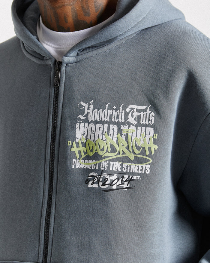 A close-up of a person wearing a heather gray zip-up hoodie with a graphic print on the chest. The graphic features the text 'Hoodrich Fils World Tour' in white and green, with 'Product of the Streets' and '2024' below.