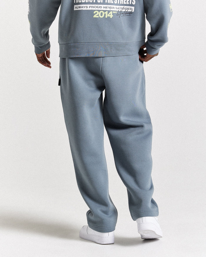 A person shown from the back wearing a matching dusty blue sweatshirt and sweatpants set with white sneakers, photographed against a plain light gray studio background.
